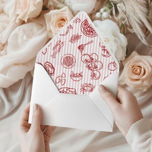May include: A white envelope held open, revealing a patterned interior with red line drawings of Italian food items like pizza, pasta, and tomatoes. The background features soft pink and white flowers.
