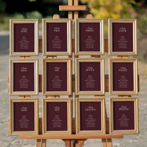Peut inclure: Un chevalet en bois présente douze cartons de table encadrés. Chaque carton doré a un fond bordeaux avec du texte blanc indiquant les numéros de table et les noms des invités. Idéal pour les mariages ou événements.