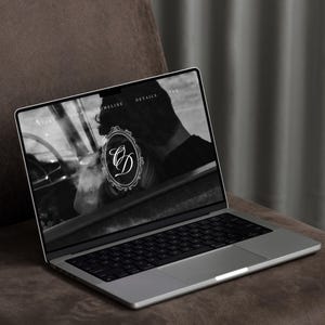 May include: A silver laptop displaying a website with a black and white image and the letters "CD" in a decorative oval frame. The laptop is on a brown surface, with a dark gray curtain in the background.