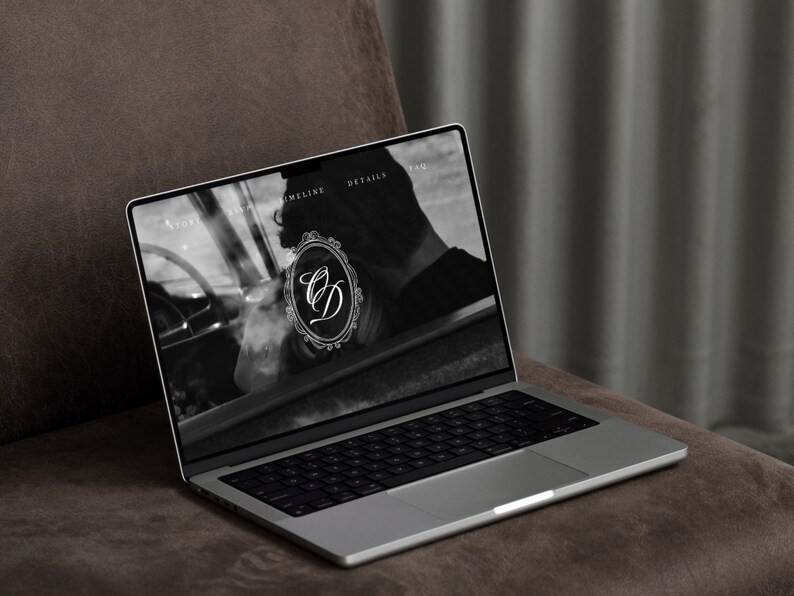 May include: A silver laptop displaying a website with a black and white image and the letters "CD" in a decorative oval frame. The laptop is on a brown surface, with a dark gray curtain in the background.