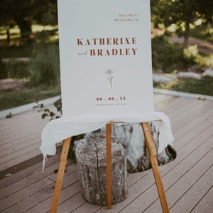 May include: A white wedding sign with brown text announcing the wedding of Katherine and Bradley on June 6, 2023 at Hardwood Creek, Lake Austin.