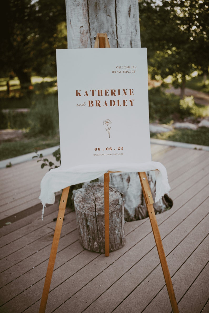 May include: A white wedding sign with brown text announcing the wedding of Katherine and Bradley on June 6, 2023 at Hardwood Creek, Lake Austin.