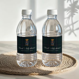 Peut inclure: Deux bouteilles d'eau en plastique transparent avec des bouchons blancs. Chaque bouteille porte une étiquette avec une illustration d'ours en peluche et le texte "Sarah's baby shower September 2, 2025" sur un fond à carreaux vert foncé. Les bouteilles sont sur un dessous de plat rond et tressé.