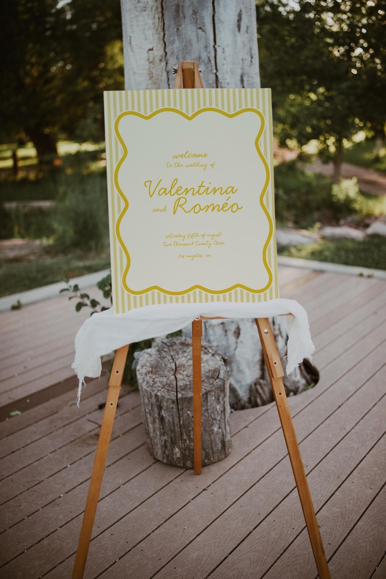 May include: A yellow and white striped sign with a white border and yellow text that reads "Welcome to the wedding of Valentina and Romeo" on a wooden easel.