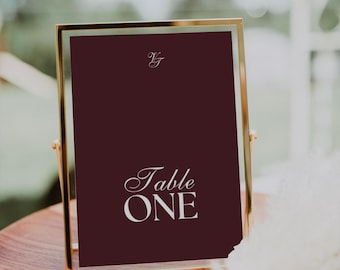 May include: A burgundy table number sign in a gold frame. The sign reads "Table ONE" in elegant white script. The top of the sign has the initials "VJ" in a delicate font. The sign is on a wooden surface.