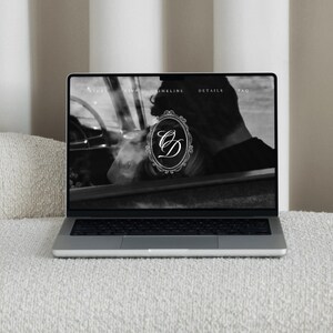 May include: A laptop displaying a website with a black and white image of a person in a car. The website's logo is a monogram within a decorative frame. The laptop is on a white textured surface.
