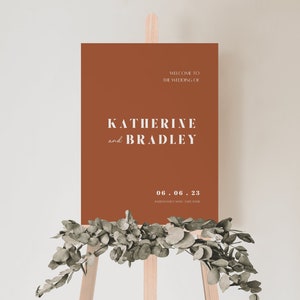 May include: A terracotta-colored welcome sign with white text announcing the wedding of Katherine and Bradley on June 6, 2023. The sign is displayed on a wooden easel with greenery.