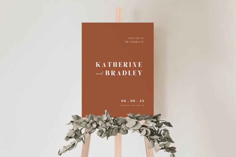 May include: A terracotta-colored welcome sign with white text announcing the wedding of Katherine and Bradley on June 6, 2023. The sign is displayed on a wooden easel with greenery.