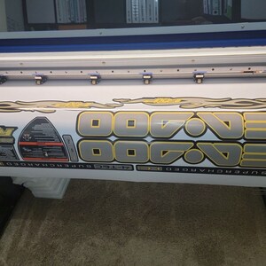May include: Close-up view of a large format printer printing a set of gray and gold vinyl decals. The decals feature the repeated number '700' with flames and other graphic elements.  Additional smaller decals include logos and text such as 'Sea-Doo' and 'Supercharged'. The decals appear to be designed for a watercraft or similar vehicle.