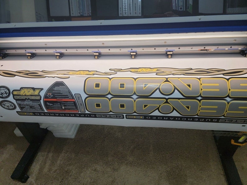 May include: Close-up view of a large format printer printing a set of gray and gold vinyl decals. The decals feature the repeated number '700' with flames and other graphic elements.  Additional smaller decals include logos and text such as 'Sea-Doo' and 'Supercharged'. The decals appear to be designed for a watercraft or similar vehicle.
