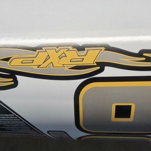 May include: Close-up view of a graphic decal featuring a stylized flame design incorporating the letters 'RXP' in gray and yellow.  Additional gray and yellow geometric shapes are present. The decal is likely intended for a vehicle or similar application. The color scheme is primarily gray, black, and yellow on a white background.