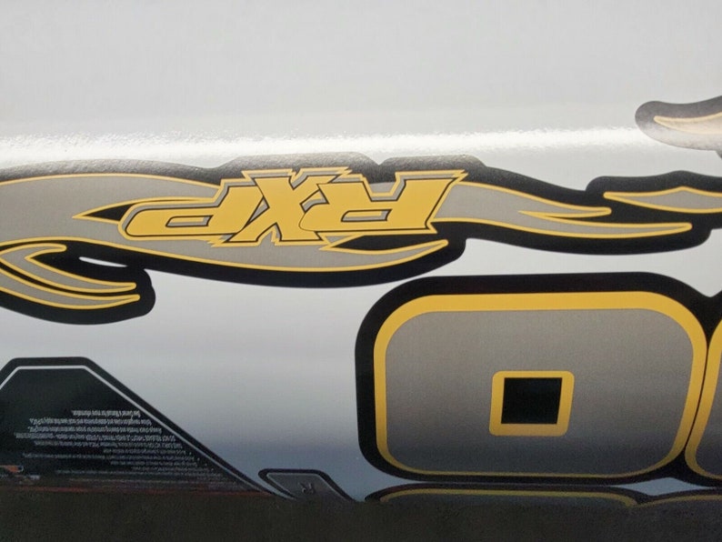 May include: Close-up view of a graphic decal featuring a stylized flame design incorporating the letters 'RXP' in gray and yellow.  Additional gray and yellow geometric shapes are present. The decal is likely intended for a vehicle or similar application. The color scheme is primarily gray, black, and yellow on a white background.