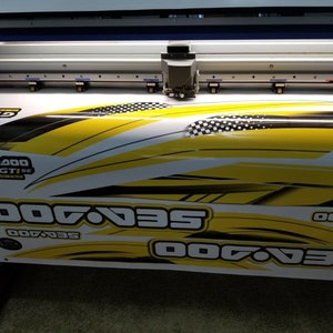 May include: A large format printer printing a yellow and black graphic design with the text "Sea-Doo" repeated multiple times. The design features a checkered pattern and black and white lines.