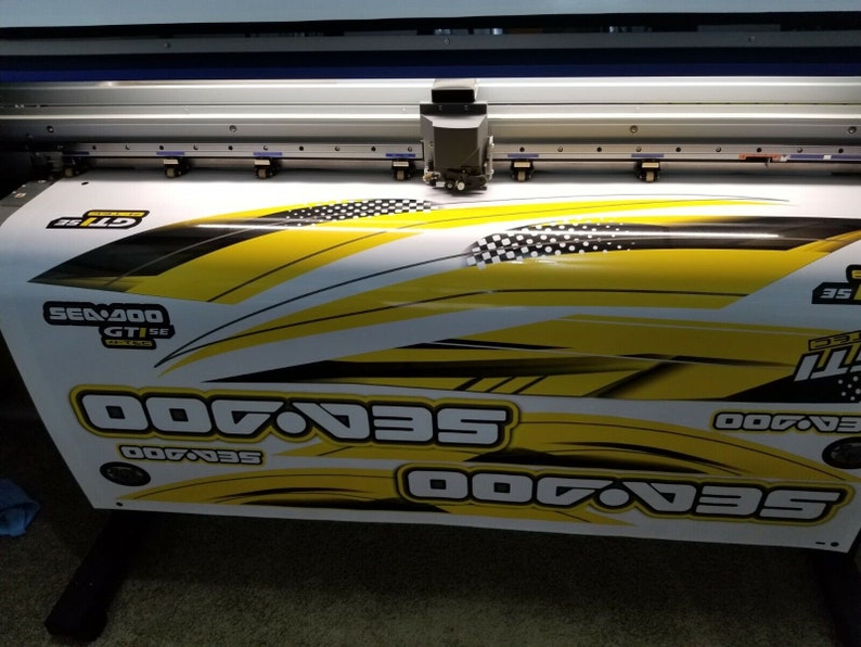May include: A large format printer printing a yellow and black graphic design with the text "Sea-Doo" repeated multiple times. The design features a checkered pattern and black and white lines.