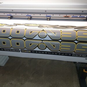 May include: Close-up view of a large format printer producing a vinyl decal. The decal features the repeated text 'SeaDoo 7-Tec Supercharged' in gray and gold lettering with a stylized flame design. The decal is printed on a white vinyl sheet and is partially rolled up on the printer. The printer is a large industrial machine with a flatbed design.