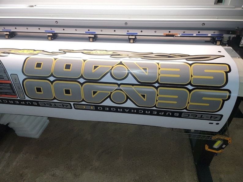 May include: Close-up view of a large format printer producing a vinyl decal. The decal features the repeated text 'SeaDoo 7-Tec Supercharged' in gray and gold lettering with a stylized flame design. The decal is printed on a white vinyl sheet and is partially rolled up on the printer. The printer is a large industrial machine with a flatbed design.