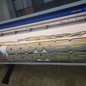 May include: Close-up view of a large-format printer producing a vinyl decal. The decal features the repeated text 'SeaDoo' in a stylized, metallic gray and gold font with yellow flames.  The decal also includes additional smaller graphics and text. The printer is a wide-format inkjet device with visible print heads and rollers.
