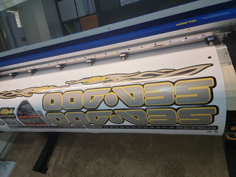 May include: Close-up view of a large-format printer producing a vinyl decal. The decal features the repeated text 'SeaDoo' in a stylized, metallic gray and gold font with yellow flames.  The decal also includes additional smaller graphics and text. The printer is a wide-format inkjet device with visible print heads and rollers.