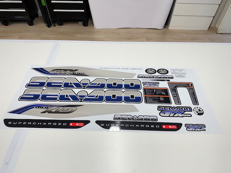 Może przedstawiać: Biała arkusz naklejek do skutera wodnego Sea-Doo GTX 2006 Limited. Naklejki są niebieskie, srebrne i czarne z nadrukiem "Sea-Doo GTX" i "Supercharged".