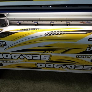 May include: A close-up of a large format printer printing a yellow and black Sea-Doo GTI SE decal. The decal features the Sea-Doo logo and the text "Sea-Doo GTI SE".