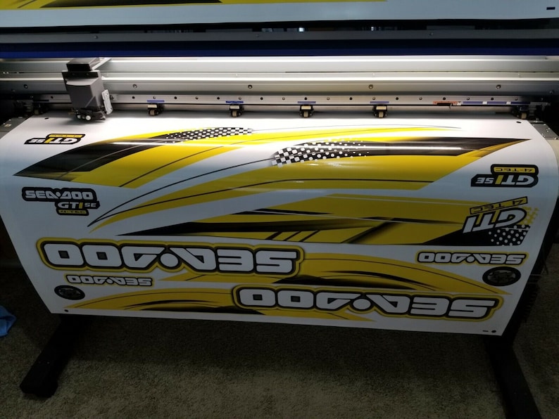 May include: A close-up of a large format printer printing a yellow and black Sea-Doo GTI SE decal. The decal features the Sea-Doo logo and the text "Sea-Doo GTI SE".
