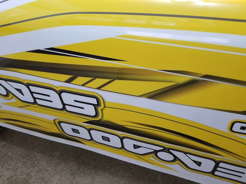 May include: Close-up of a yellow and white graphic design with the word "Sea-Doo" in black and white lettering.