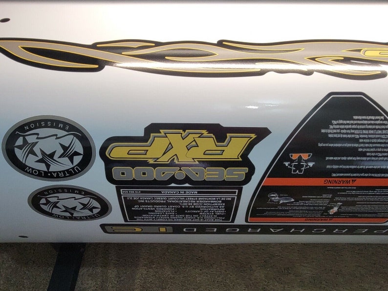 May include: Close-up view of Sea-Doo 700 decals and labels.  Features include a gray and yellow flame design, two Ultra-Low Emission circular logos, a rectangular Sea-Doo 700 label with yellow lettering, and a triangular warning label with safety information.  All decals are on a white background. The labels include text such as 'Ultra-Low Emission', 'Sea-Doo 700', and 'Warning'. The overall theme is watercraft or personal watercraft graphics and labels.