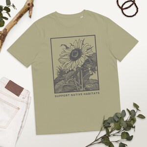 Op de afbeelding: Een lichtgroen T-shirt met een zwart-witte illustratie van een zonnebloem in een kader. De tekst "SUPPORT NATIVE HABITATS" staat onder de afbeelding.