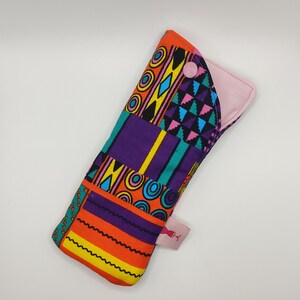 Könnte beinhalten: Eine bunte Stoff-Menstruationseinlage mit geometrischem Muster und einem rosa Knopf. Die Einlage ist in der Mitte gefaltet und hat ein rosa Futter.