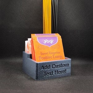 May include: A gray 3D printed tea bag and stirrer holder with the text "Add Custom Text Here!" and a stack of Yogi Sweet Tangerine Positive Energy tea bags. The holder also contains a bundle of black stirrers and two yellow straws.