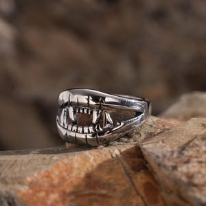 May include: A silver-colored ring with a detailed design of open jaws and sharp teeth. The ring is set against a natural, textured background, highlighting its metallic sheen and intricate craftsmanship. The ring's design evokes a sense of boldness and unique style.
