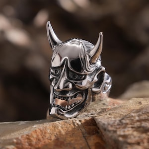 May include: A silver ring with a detailed, textured design of a Japanese Oni mask. The mask has horns, a wide mouth, and sharp teeth.
