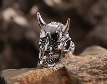 Puede incluir: Un anillo de plata con un diseño detallado y texturizado de una máscara Oni japonesa. La máscara tiene cuernos, una boca ancha y dientes afilados.