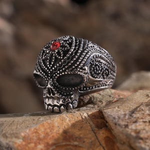 May include: A silver skull ring with intricate details and a red gemstone in the centre of the forehead.