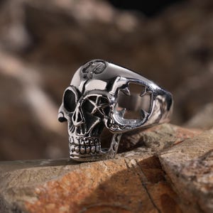 May include: A silver skull ring with intricate details. The skull has a cross on its forehead and a cut-out design on the side. The ring is set on a rough, brown rock.