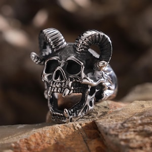 May include: A silver skull ring with horns. The skull has a detailed design with an open mouth and sharp teeth.
