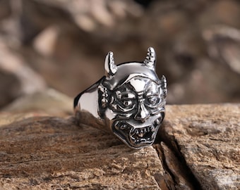 Puede incluir: Un anillo de plata con un diseño detallado de la cara de un demonio. El anillo está colocado sobre un fondo de madera rústico.