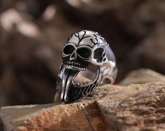 May include: A silver skull ring with an open mouth and detailed design. The ring is set against a brown, textured background.