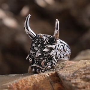 May include: A silver-colored ring featuring a detailed oni mask design. The mask has prominent horns, sharp teeth, and intricate facial features. The ring is set against a natural, textured background.