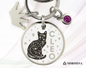 Może przedstawiać: Srebrny charm w kształcie kota z galaktycznym wzorem i wygrawerowanym imieniem "CLEO". Charm jest przymocowany do srebrnego breloka z koroną i różowym kryształem.