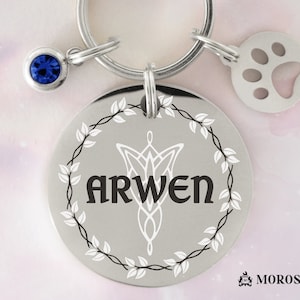 May include: Silver dog tag with a blue gem, a paw print charm, and a Celtic knot design. The tag is engraved with the name "Arwen" and a wreath of leaves.