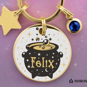 May include: Gold-toned keychain with a round charm featuring a black cauldron with the name "Felix" in gold lettering. The charm has a white background with gold stars and a blue gemstone charm.
