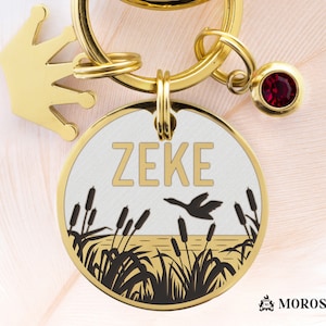 May include: Gold-tone keychain with a round tag featuring a white background, black silhouette of cattails and a bird, and the name "ZEKE" in gold lettering. The keychain also includes a small gold crown charm and a red gemstone charm.