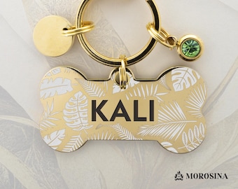 May include: Gold-colored dog tag in a bone shape, featuring the name "KALI" in black block letters. The tag is adorned with a tropical leaf pattern in white. A gold ring and charm with a green gem are attached.