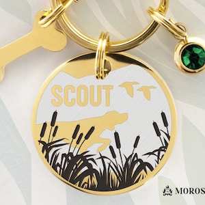 May include: Gold dog tag with a black silhouette of a dog and reeds, two flying ducks, and the word "SCOUT" in white. The tag is attached to a gold key ring with a bone charm and a green gemstone.