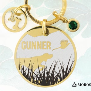 Puede incluir: Una placa de perro de oro con fondo blanco y silueta negra de un perro corriendo por la hierba alta. La placa también presenta un faisán volando sobre el perro y el nombre "GUNNER" en letras doradas. La placa está unida a un llavero de oro con un dije de hueso y un dije de piedra preciosa verde.