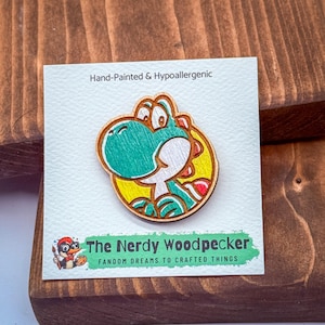 May include: A wooden pin featuring a cartoon Yoshi character with green, white, red, and yellow colors. The pin is hand-painted and hypoallergenic. The Nerdy Woodpecker logo is at the bottom.