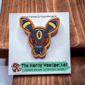 May include: A wooden pin featuring a cartoon character with black, yellow, and red details. The pin is hand-painted and hypoallergenic. The pin is on a white card with the text "The Nerdy Woodpecker" and "Fandom Dreams to Crafted Things".