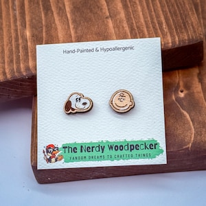 May include: Wooden stud earrings featuring Snoopy and Charlie Brown from the Peanuts comic strip. The earrings are hand-painted and hypoallergenic. The Nerdy Woodpecker logo is visible on the card.