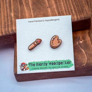May include: Hand-painted, hypoallergenic wooden earrings featuring a paintbrush and a paint palette. The earrings are on a white card with the text "Hand-Painted & Hypoallergenic" and "The Nerdy Woodpecker Fandom Dreams to Crafted Things."